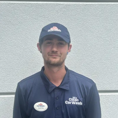 Man wearing navy blue Mr Clean Car Wash uniform and cap standing against a gray textured wall.