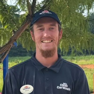 Smiling man wearing a Mr. Clean Car Wash uniform and name tag standing outdoors by a tree.