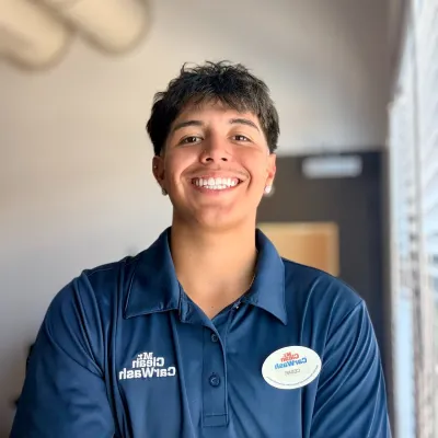 Smiling young man in a navy blue Mr. Clean Car Wash uniform with crossed arms indoors near window blinds.