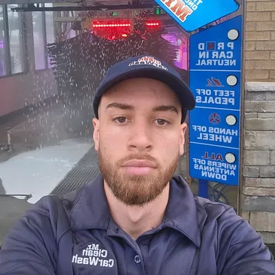 Young man in Mr. Clean Car Wash uniform takes selfie inside car wash with blue and red signage in background.