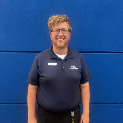 Smiling man with glasses wearing a navy CarWash polo shirt standing against a blue wall with gravel beside him