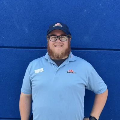 Smiling man in Mr. Clean Car Wash uniform standing against a deep blue wall with hands on hips.