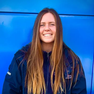 Smiling young woman with long hair wearing a navy blue jacket standing against a bright blue wall.