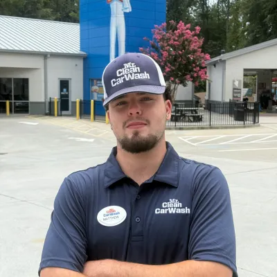 Employee in uniform standing confidently outside Mr. Clean Carwash location with billboard of Mr. Clean mascot