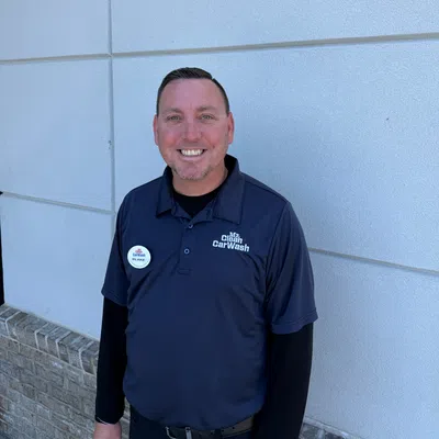 Smiling car wash employee wearing navy blue Mr. Clean CarWash uniform and name badge standing against white wall.