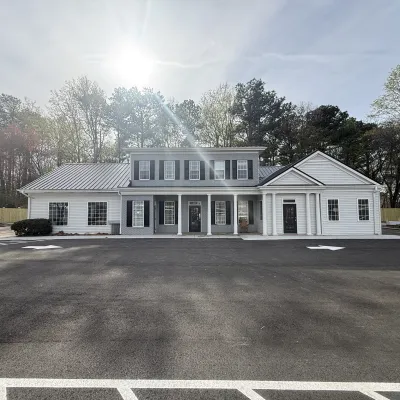 White two-story building with black shutters surrounded by trees and a large empty parking lot under a sunny sky