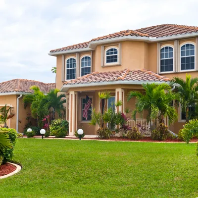 Florida house with red tiled roof surrounded by green lawn and tropical plants under cloudy sky
