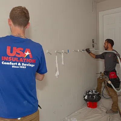 Two workers installing spray foam insulation inside a basement with USA Insulation shirt visible.