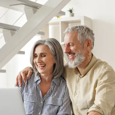 Happy mature couple sitting on couch using laptop together under modern staircase in bright living room