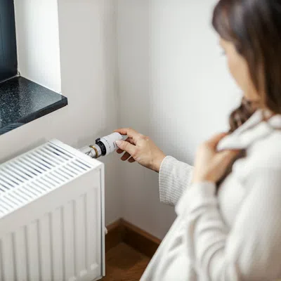 Woman adjusting the temperature knob on a white radiator heater next to a window in a cozy room.