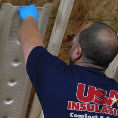 Worker wearing blue gloves installing insulation panels on an attic ceiling with exposed wooden beams.