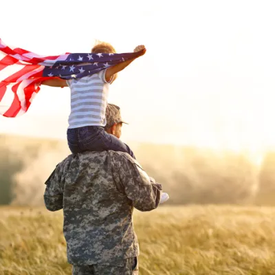 A soldier carries a child with an American flag in a sunlit, open field, symbolizing patriotism and family.