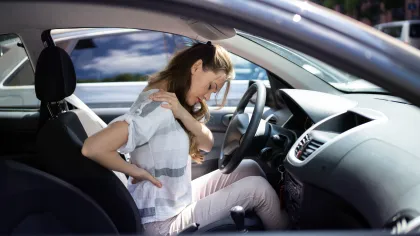 Woman sitting in a car holding her back and shoulder in pain, indicating discomfort or injury.