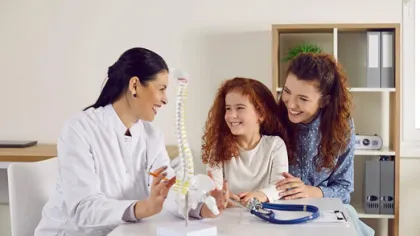 Doctor explaining spine model to a smiling child and her mother during a medical consultation.