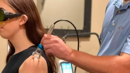 Woman wearing protective glasses receiving laser therapy on shoulder from a technician in clinical setting.
