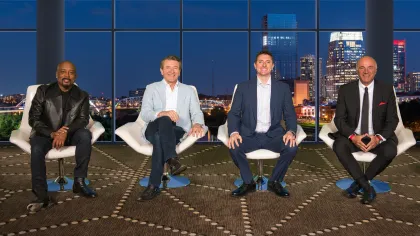 Four men in business attire sitting on white chairs with a city skyline at night in the background.