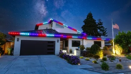 Modern house decorated with red, white, and blue LED lights under a starry night sky with American flag and landscaping.