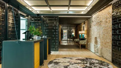 Modern hotel lobby with yellow reception desks, exposed brick wall, patterned rug, and decorative chairs.