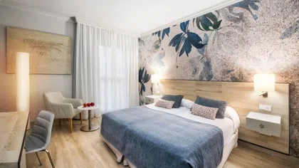 Modern hotel room with a queen bed, blue bedding, floral accent wall, and cozy seating area.