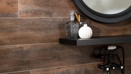 Bathroom wall with wood-look tiles, a round mirror, black faucet, floating shelf, and decorative items.