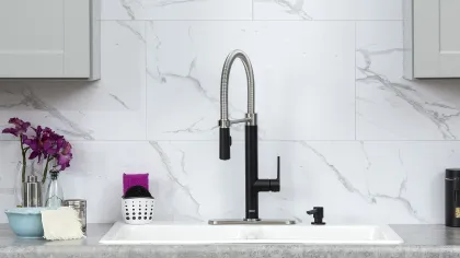 Modern kitchen sink with black and silver faucet against white marble backsplash and gray cabinetry.