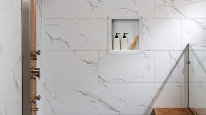 Modern shower area with marble tiles, rain showerhead, built-in niche with bottles, and wood bench with towels