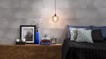 Modern bedroom with gray tiled wall, wooden bench, decorative items, and bed with patterned pillows and black cover.