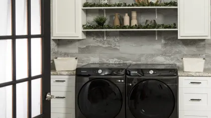 Modern laundry room with black Samsung washer and dryer, white cabinets, gray tile wall, and decorative shelves.
