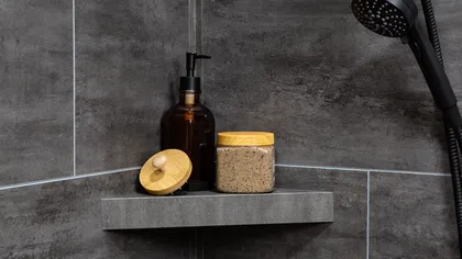 Modern shower with dark grey tiles, black showerhead, amber soap dispenser, and scrub jar on a corner shelf.