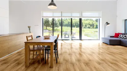 Bright modern dining and living area with wooden floors, large windows, natural light, and outdoor greenery.
