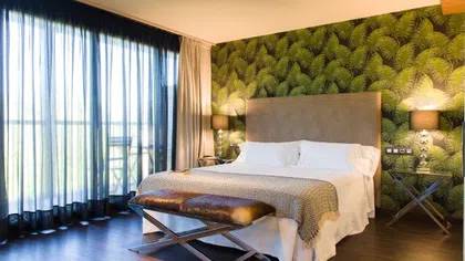 Modern bedroom with large bed, tropical leaf wallpaper, sheer curtains, and wooden floor accents.
