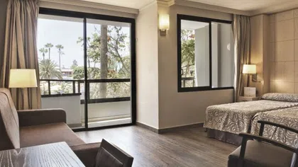 Bright hotel room with two beds, sitting area, wooden floor, large windows, and balcony with outdoor view.