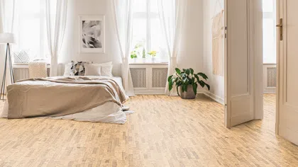 Bright bedroom with large windows, white curtains, beige bedding, wooden floor, and green potted plant