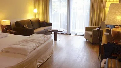 Cozy hotel room with a bed, sofa, armchair, wooden floor, and large windows with sheer curtains letting in natural light