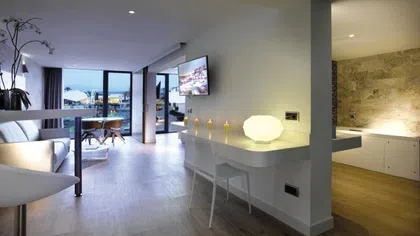 Modern studio apartment with open layout, white furniture, wooden floors, and large windows overlooking cityscape at dusk.