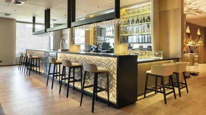 Modern bar area with high stools, illuminated patterned counter, wooden floors, and shelves with bottles and glasses.