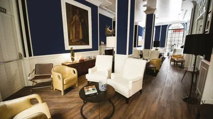 Elegant lounge with white and tan chairs, wooden floors, blue walls, and classic paintings in a spacious room.