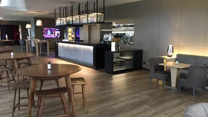 Modern lounge area with wooden tables, padded chairs, sleek bar counter, and ambient lighting in a spacious room