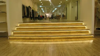 Modern retail store interior with wooden flooring, illuminated stairs, and clothing displays along white walls.