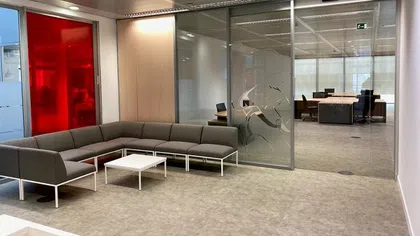 Modern office waiting area with gray sectional sofa, white tables, red tinted glass wall, and glass office partition.