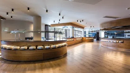 Modern empty buffet area with circular wooden counters, glass sneeze guards, and natural lighting on wooden floors.