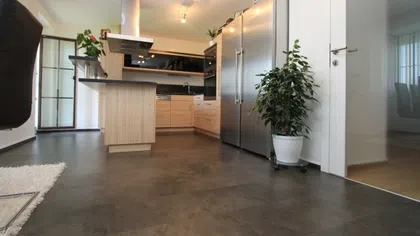 Modern kitchen with wooden cabinets, stainless steel refrigerator, island, and potted plant on polished floor.