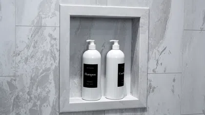 White shampoo and conditioner bottles with black labels in a marble shower niche with gray veining.