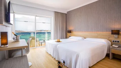 Bright hotel room with two beds, wooden floors, balcony overlooking the sea, and modern decor.