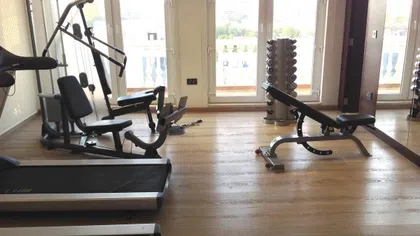 Home gym with treadmill, weight machine, adjustable bench, free weights, and wooden flooring in bright room.