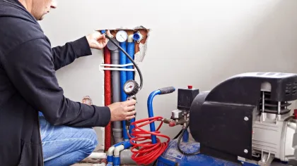 Technician checking water pressure gauges on pipes connected to a blue air compressor in a wall cavity.