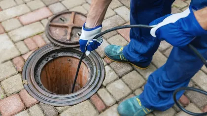 Worker wearing gloves and boots inspecting an open manhole with a cable on a brick-paved surface.