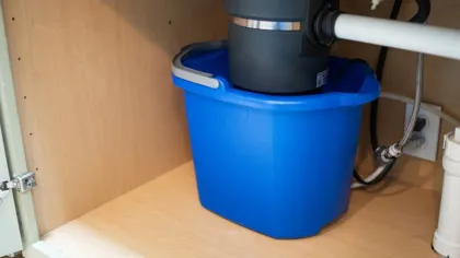 Blue bucket placed under kitchen sink pipes and garbage disposal unit inside wooden cabinet.