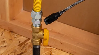 Close-up of a yellow gas valve and flexible connector pipe installed on a wooden floor and wall framing.