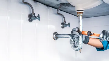 Plumber wearing gloves repairing a metal pipe underneath a sink in a tiled bathroom or kitchen setting.